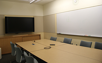 Classroom with four rectangular tables arranged into one larger rectangle. Blue chairs around the table. White board on one wall. A large television monitor mounted to the front wall.