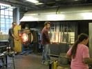 Photo of one glassblower heating glass at a furnace and another collecting hot glass at a second furnace.