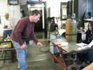 Photo of a glassblower rolling another glob of hot glass with white rods that will become a second vase.