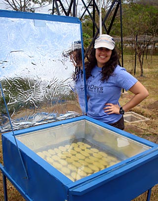 Photo of a student standing outdoors next to a box containing cookies, with clear plexiglass top and a large foil solar reflector.