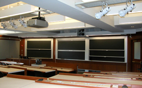 Tiered lecture hall with chalkboards and projector at the front of the room and grouped seating for students.