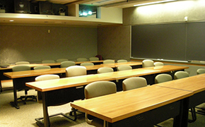 Four rows of tables with padded chairs on wheels. A/V equipment in the rear of the room and a chalkboard on the sidewall.