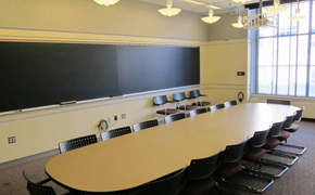 Photo shows the classroom with a long, oval seminar table surrounded by chairs and chalkboards on the walls.