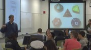 An instructor stands and speaks to a classroom of students. A dozen students are pictured, sitting around two round tables. Projector screens show a comic and a set of dice.