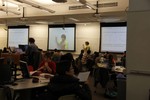 An instructor stands at a whiteboard while students sit at a round table. The instructor is filmed by a ceiling-mounted camera and the recording is live-streamed on screens around the classroom. Alternate screens show a slide.