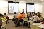An instructor writes on a whiteboard while students look on. The instructor is captured by a ceiling-mounted camera and live-streamed on screens around the classroom. 