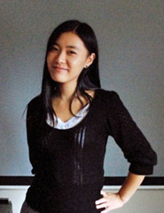 A woman with black hair in a black sweater stands in front of a blackboard. She has her hand on her hip.