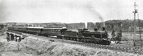 Archival black-and-white photo, side view of steam locomotive pulling several cars over a low bridge. Steam is blowing up out of the whistle.