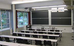 This classroom photo shows five rows of student tables, each with six chairs. At the front there is a smaller table and two chairs, and two sets of sliding chalkboards.