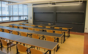 4 rows of tables and chairs facing blackboards at the front of the room. Windows line the left wall.