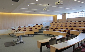 Wooden lecture tables arranged in tiers; red padded chairs on wheels behind each row, grey carpet. Windows on rear wall; lectern with monitor at front of room.