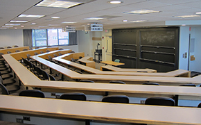 Five rows of lecture tables with grey chairs; black sliding boards at the front of the room; a window on the left wall.