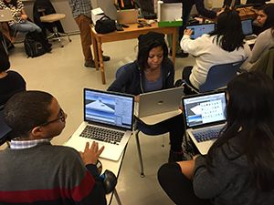 Three students in a classroom, huddled around laptops.
