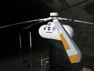 Side-view of a test helicopter in a wind tunnel with a focus on one of the blades.
