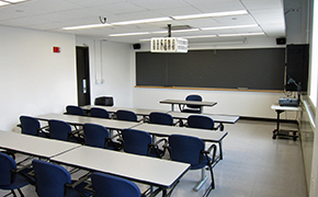 This photo shows the room the lectures were given in with rows of tables and moveable chairs for a maximum of 20 students.  There is an overhead and manual slide projector, a large chalkboard at the front and a window to the students right.