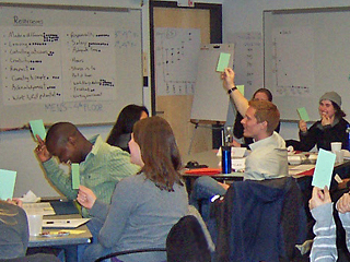 Students sitting in class raise their hands while holding green notecards.