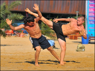 Two men engaging in capoeira martial arts on a beach. The man on the right is performing a high kick, and the man on the left has his arms extended upward and is using his foot to trip his opponent.