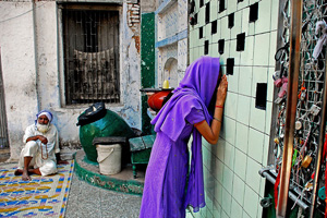 A photo of a girl looking through a window in Ahmedabad. Behind her is an old man, sitting on the ground, and a plastic bucket. By her right arm is a grating covered in padlocks.