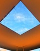 A photo of a ceiling with a square opening revealing blue skies and clouds above.