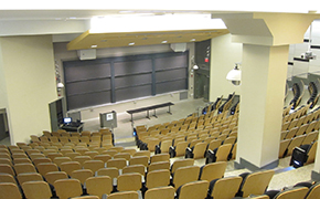 View of lecture hall from back of the classroom. Tiered seating. Chalkboards and lectern at front of room.
