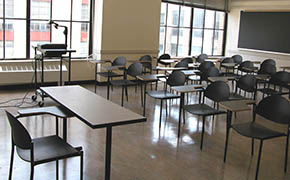 View from front of classroom. Five rows of tablet desks positioned in the center of classroom. Wall of windows to the left of desks.