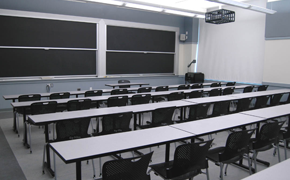 A classroom with four long tables spanning the classroom, chairs behind each table, and four sliding chalkboards at the front.