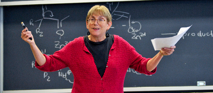 A teacher in a red sweater standing in front of a blackboard, gesturing to her audience.