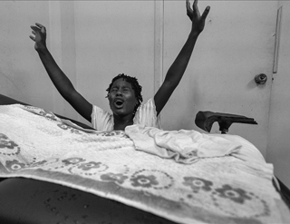 A black woman standing behind a simple hospital bed. Her arms are outstretched, her eyes are close, and her mouth is open, as if she is screaming.