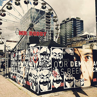 A photo of a ferris wheel with the words, "Mr. Robot" on it. In the foreground are two walls plastered with posters from the TV series.