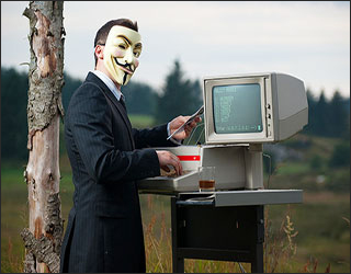 A man in a suit wears a Guy Fawkes mask on his face while standing next to a computer.