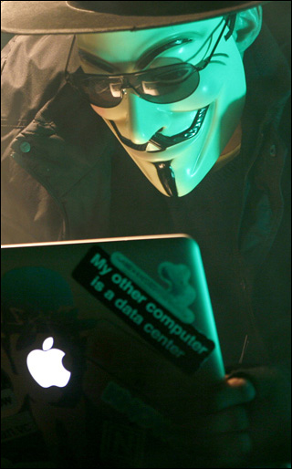 Disguised by the mask of Guy Fawkes, the Anonymous Hacker peers at his laptop screen through black glasses.