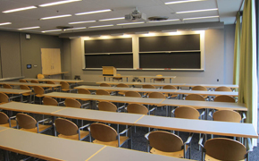 A photograph of a classroom with multiple blackboards in the front of the room. Students sat at long tables.