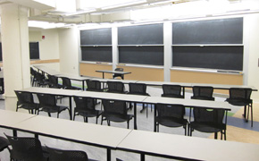 A large classroom with three rows of tables and chairs facing three chalkboards.