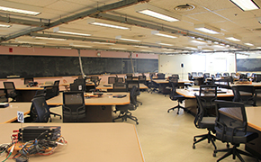 Classroom with 13 square tables visible. Four laptops are on the table closest to the viewer. Four black chairs on wheels are arranged around each table. A blackboard lines one wall.