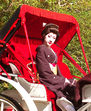 A small, keen-featured man, clothed in a kimono, and wearing powder on his face and rouge on his lips, sits in a carriage.