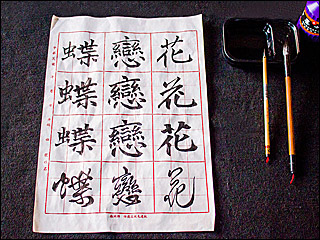Three Chinese characters in four different calligraphy scripts written on calligraphy grid paper, with two brushes, a bottle of ink, and an inkstone at the side.
