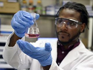 A young male, wearing goggles and gloves, observes  purple liquid in a beaker.