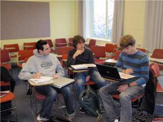 Three students sitting at desks, reading to each other from textbooks.