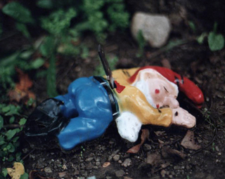 Ceramic figurine with red cap face down in the dirt with a knife in his back.