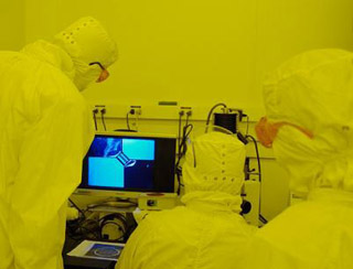 People in cleanroom suits view magnified wafer pattern on screen.