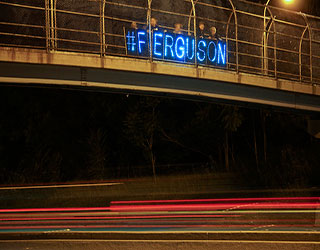 Protesters stand on a highway overpass with a lit up sign that reads "#FERGUSON".