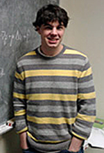 A young man with black hair is standing beside a chalkboard. He is wearing a sweater with grey and yellow horizontal stripes. His hands are in his pockets.