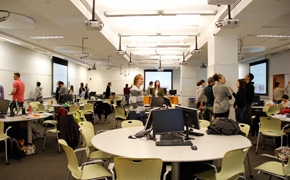 Groups of students work at white boards around the classroom; three projector screens display the same math problem. The center of the room is filled with round tables that are surrounded with chairs. Numerous projectors and cameras are mounted from the ceiling.