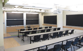 A classroom with four long tables spanning the classroom, chairs behind each table, and two sliding chalkboards at the front.