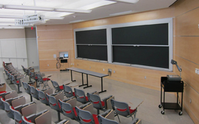 Classroom with sliding chalkboards, two narrow tables in front of the chalkboards, three rows of tiered seating with tablet desks, A/V equipment, and an overhead projector.