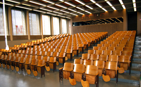 Lecture hall viewed from the front of the room facing toward the rear of the room. Windows line the left side of the room. Fifteen rows of seats with arm tablets are visible.