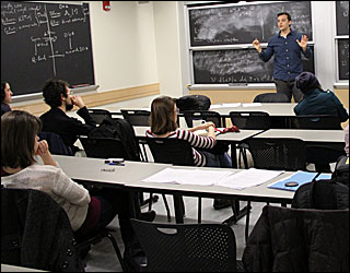 Male student in blue dress shirt in fron of black board and speaking to a small group of students.