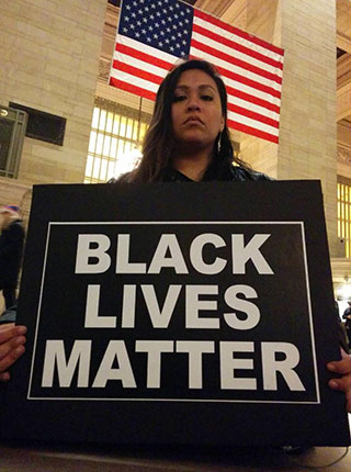 A solemn-faced brown-skinned woman, seated beneath the American flag, holds a sign with the text: Black Lives Matter.