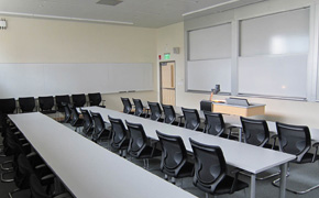 Photo of a high-ceiling classroom with long row tables for students and whiteboards on both the front and side walls.