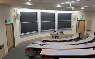 Classroom with 5 tired and curved rows of tables. Red chairs positioned behind the tables. Three sliding chalkboards at the front of the room.